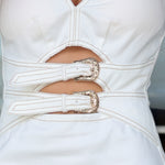 Close-up of a white sleeveless dress with cutouts at the waist and two decorative silver-tone buckles, worn by a model.