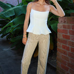 A woman stands outdoors wearing a white spaghetti-strap peplum top, beige zebra-print cropped pants, and white sneakers, posed in front of a brick wall and large green tropical leaves.