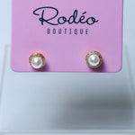 A pair of stud earrings with white pearls and gold-toned halo settings, displayed on a pink Rodéo Boutique card against a light gray background.