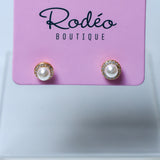 A pair of stud earrings with white pearls and gold-toned halo settings, displayed on a pink Rodéo Boutique card against a light gray background.