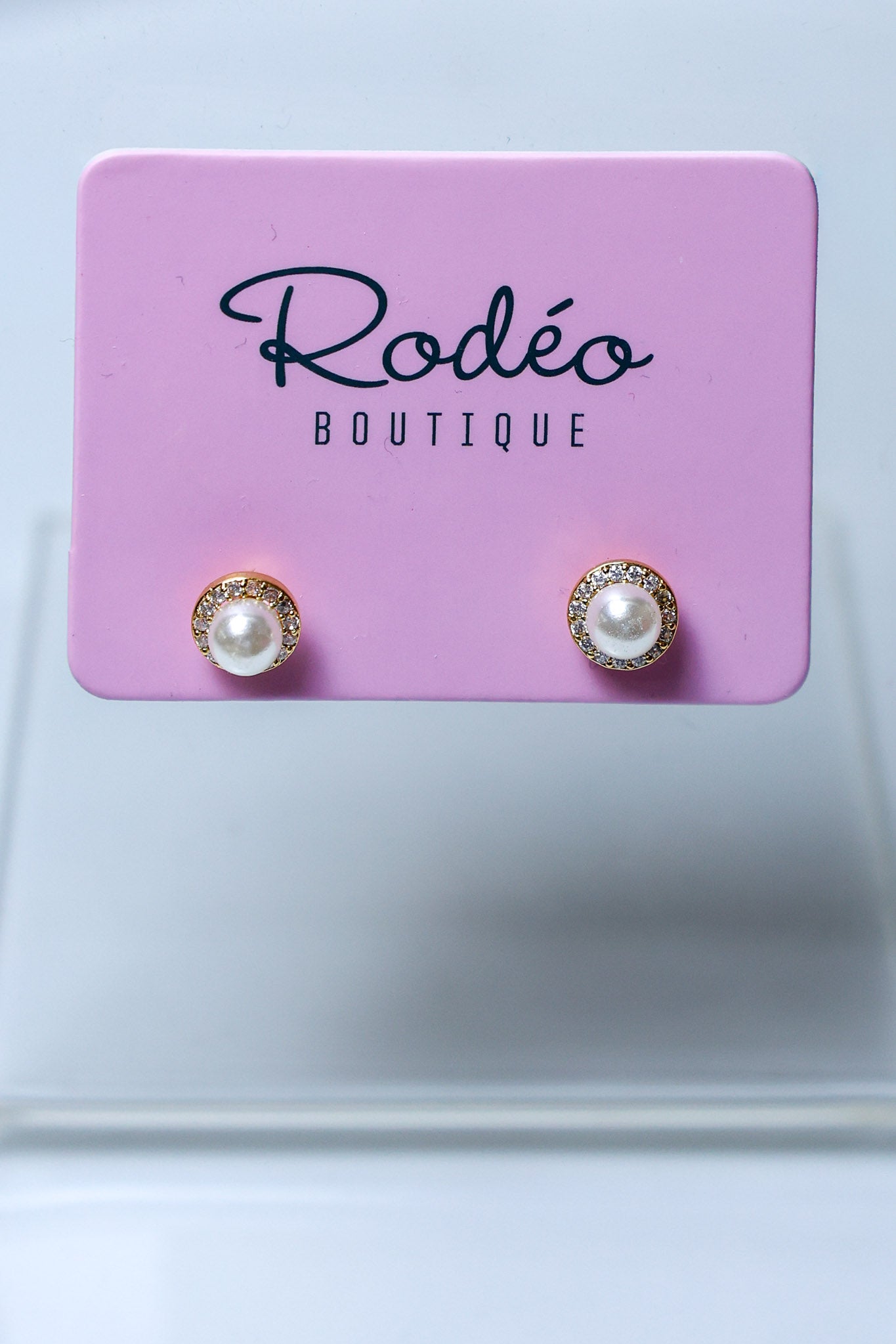 A pair of stud earrings with white pearls and gold-toned halo settings, displayed on a pink Rodéo Boutique card against a light gray background.