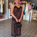 A woman stands in a boutique wearing a brown sheer maxi slip dress with black lace trim, holding a black handbag. The background shows clothing racks and a wooden floor.