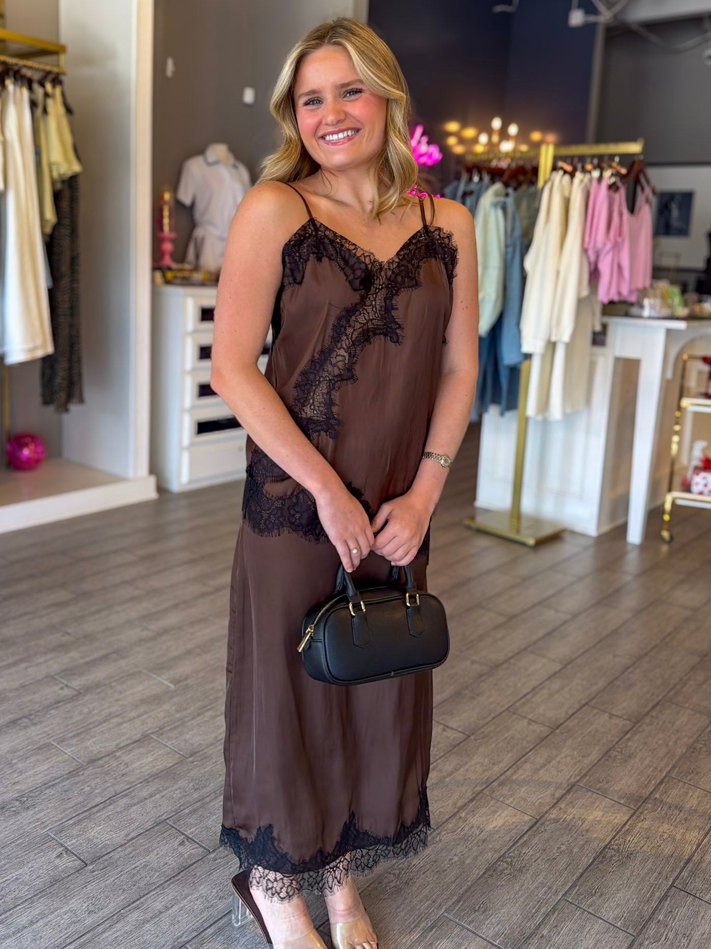A woman stands in a boutique wearing a brown sheer maxi slip dress with black lace trim, holding a black handbag. The background shows clothing racks and a wooden floor.