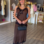 A woman stands in a clothing boutique wearing a brown sheer maxi dress with black lace trim, holding a small dark blue handbag.