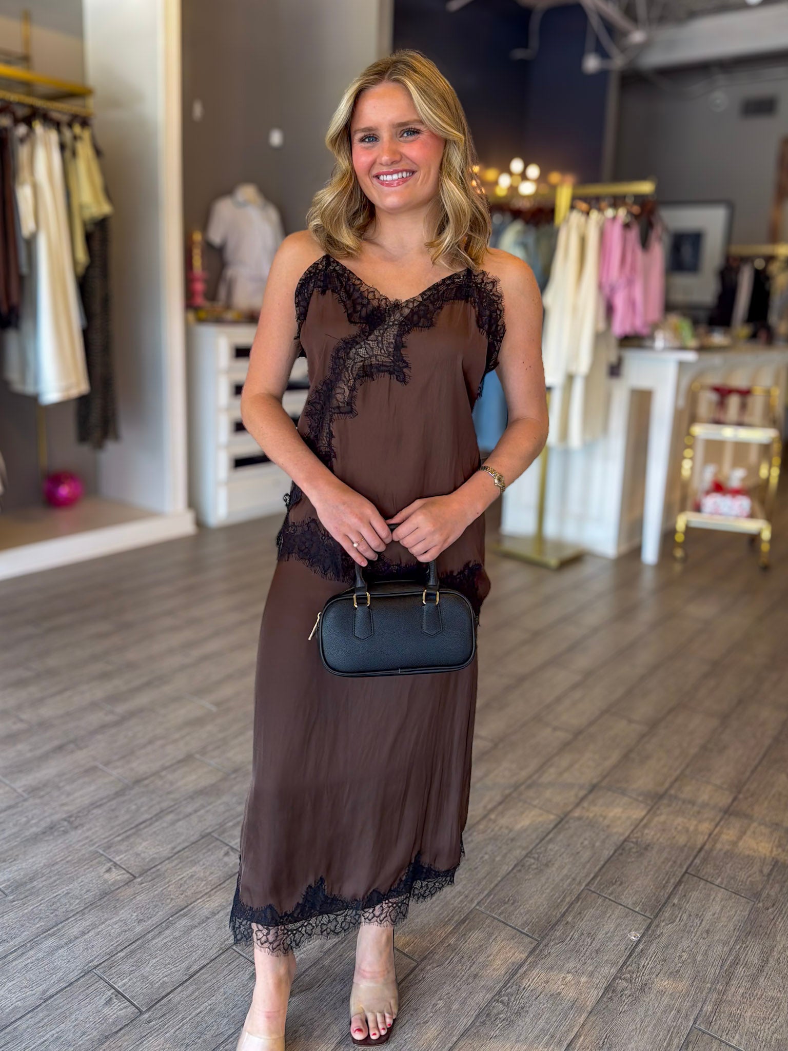 A woman stands in a clothing boutique wearing a brown sheer maxi dress with black lace trim, holding a small dark blue handbag.
