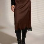 A model wearing a brown satin midi skirt with a black lace hem, styled with a black long-sleeve top and black pointed-toe ankle boots.