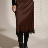 A model wearing a brown satin midi skirt with a black lace hem, styled with a black long-sleeve top and black pointed-toe ankle boots.