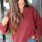A woman wearing a long-sleeve quarter-zip pullover in a heathered burgundy red, paired with blue jeans, standing in front of a glass door.