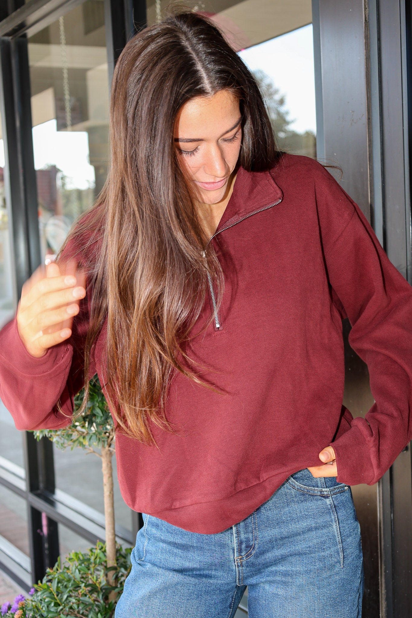 A woman wearing a long-sleeve quarter-zip pullover in a heathered burgundy red, paired with blue jeans, standing in front of a glass door.