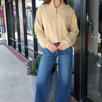 A woman stands on a sidewalk in front of a storefront, wearing a beige quarter-zip long-sleeve top, wide-leg blue jeans, and red sneakers.
