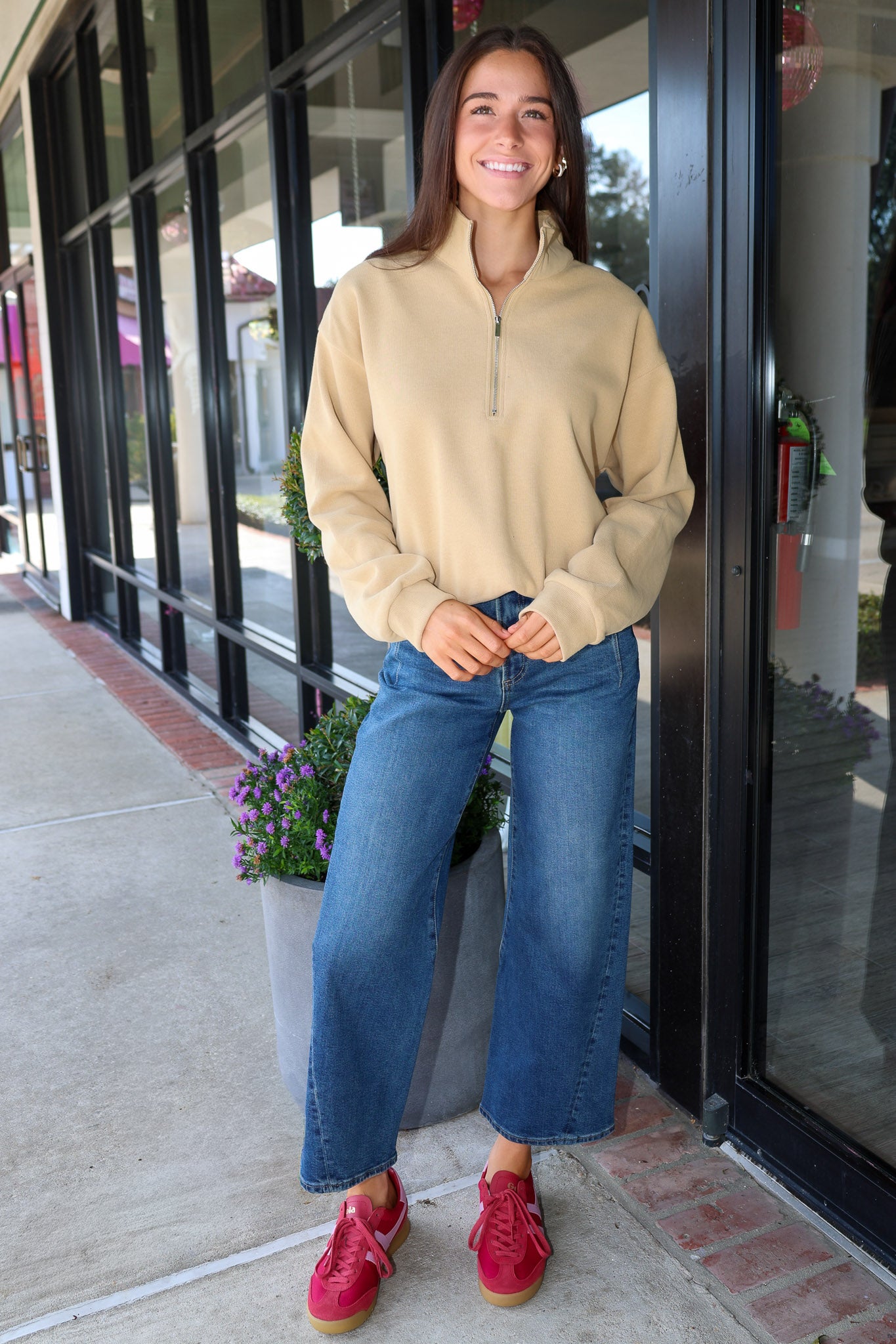 A woman stands on a sidewalk in front of a storefront, wearing a beige quarter-zip long-sleeve top, wide-leg blue jeans, and red sneakers.