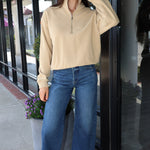 A woman stands on a sidewalk in front of a storefront, wearing a beige long-sleeve quarter-zip pullover, blue wide-leg cropped jeans, and red sneakers.