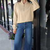 A woman stands on a sidewalk in front of a storefront, wearing a beige long-sleeve quarter-zip pullover, blue wide-leg cropped jeans, and red sneakers.
