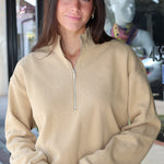 A woman wearing a beige quarter-zip pullover sweatshirt and blue jeans, standing in front of a storefront window.