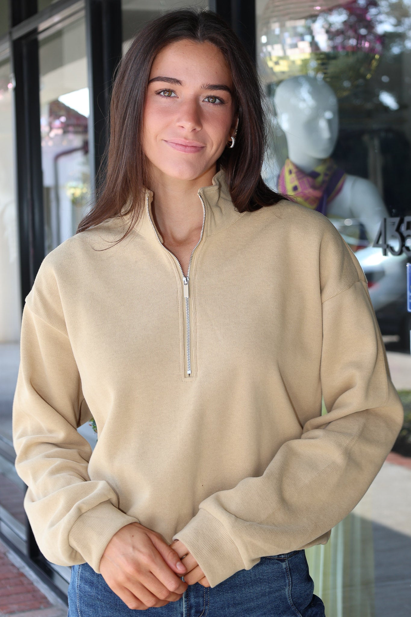 A woman wearing a beige quarter-zip pullover sweatshirt and blue jeans, standing in front of a storefront window.