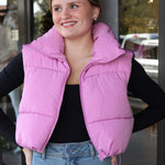 Person wearing a pink puffer vest and blue jeans standing outside a building.