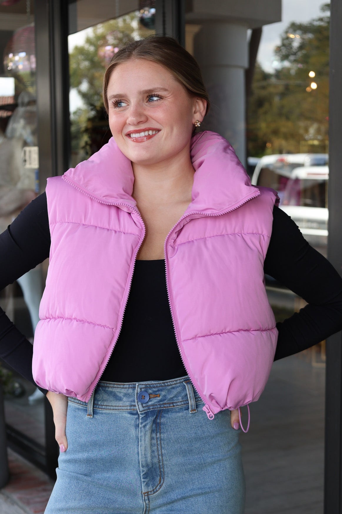 Person wearing a pink puffer vest and blue jeans standing outside a building.