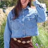 Person wearing a denim jacket and brown skirt in an outdoor setting with grass and flowers.
