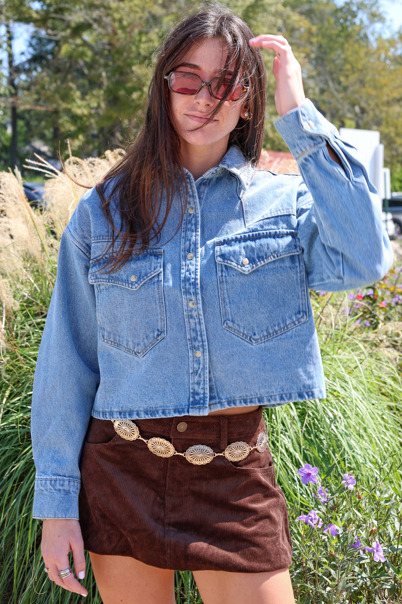 Person wearing a denim jacket and brown skirt in an outdoor setting with grass and flowers.