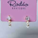 A pair of gold hoop earrings with clear round-cut gemstones and white pearl drops, displayed on a pink Rodéo Boutique card.