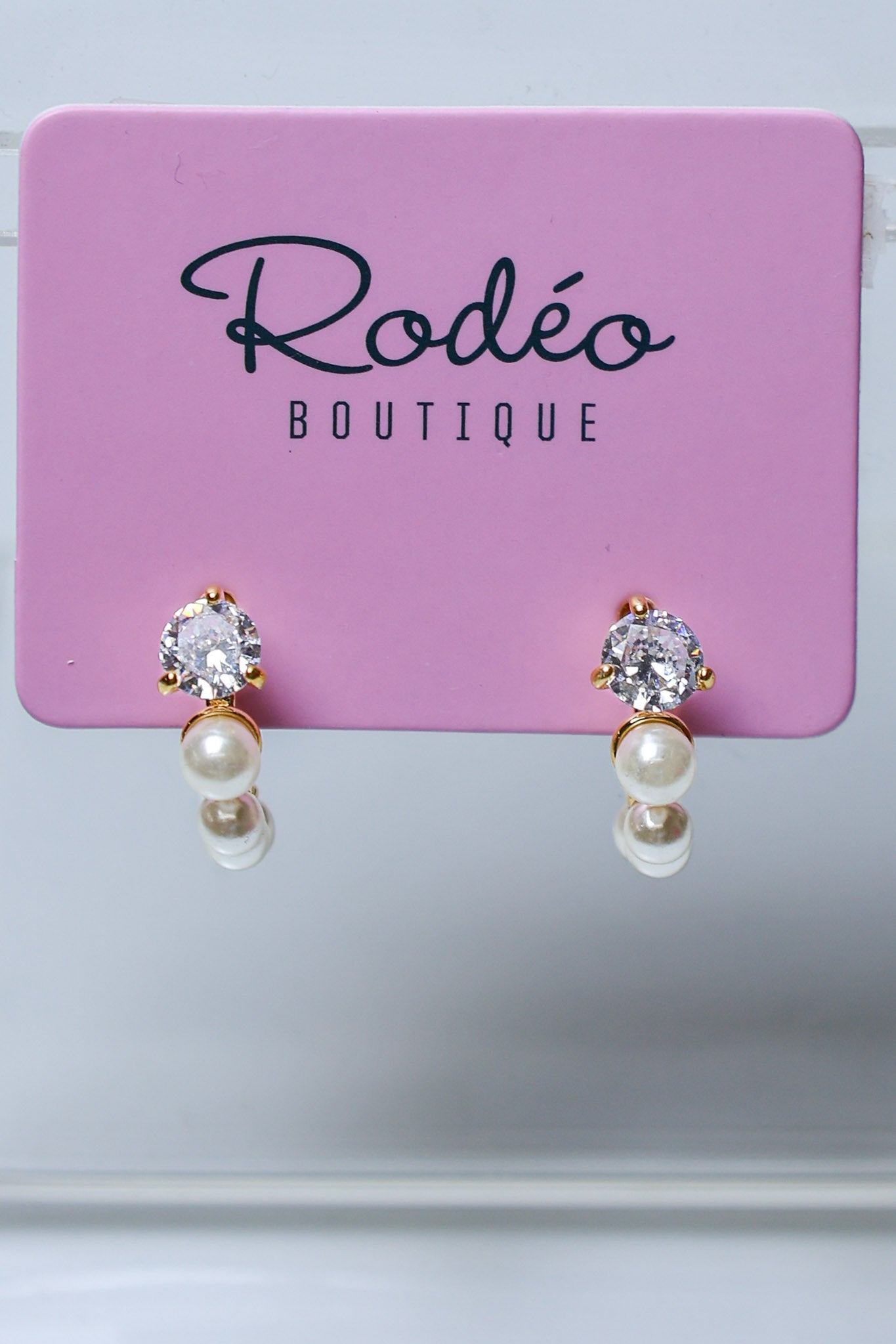 A pair of gold hoop earrings with clear round-cut gemstones and white pearl drops, displayed on a pink Rodéo Boutique card.