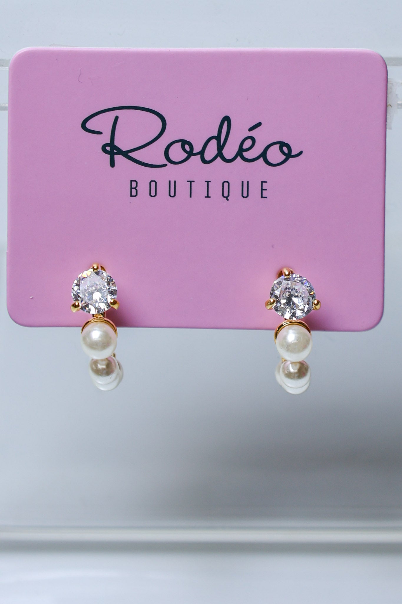 A pair of gold hoop earrings with clear round-cut gemstones and white pearl drops, displayed on a pink Rodéo Boutique card.