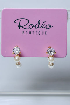 A pair of gold hoop earrings with clear round-cut gemstones and white pearl drops, displayed on a pink Rodéo Boutique card.