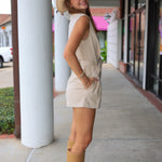 A woman posing Rodéo Boutique wearing a beige sleeveless romper, a wide-brimmed straw hat, and tan knee-high boots.