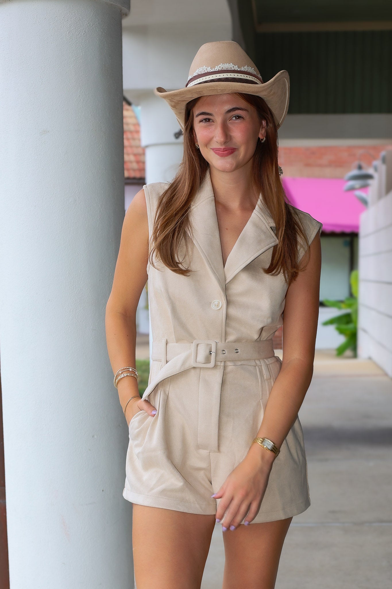 A woman wearing a beige sleeveless belted romper and a matching cowboy hat, standing in a covered walkway near Rodéo Boutique.