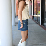 Woman wearing a beige sleeveless top, denim skirt, and white cowboy boots standing on a sidewalk.