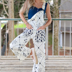 Woman wearing a white floral overalls with a blue top, standing on a wooden deck by Free People