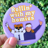 A close-up of a hand holding a round purple button badge with the text 'Rollin' with my homies' and an illustrated tiger and football player, against a blurred green outdoor background.