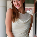 A smiling young woman with sunglasses on her head stands in a porch setting wearing a sleeveless cream cowl-neck top and light khaki pants.