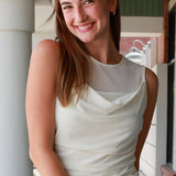 A smiling young woman with sunglasses on her head stands in a porch setting wearing a sleeveless cream cowl-neck top and light khaki pants.