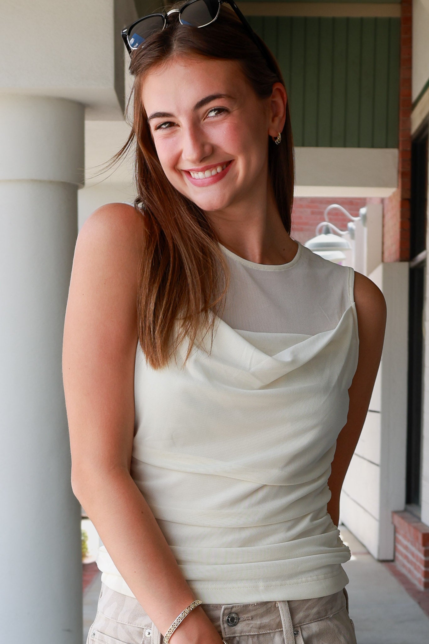 A smiling young woman with sunglasses on her head stands in a porch setting wearing a sleeveless cream cowl-neck top and light khaki pants.