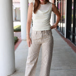 A woman stands on a covered walkway wearing a sleeveless cream top and beige patterned cropped pants with white heeled sandals. She has sunglasses on her head and gold bracelets on her wrist.