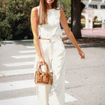 A woman walking outdoors on a sunny street crosswalk wearing a white sleeveless top and matching wide-leg trousers, holding a brown leather handbag.