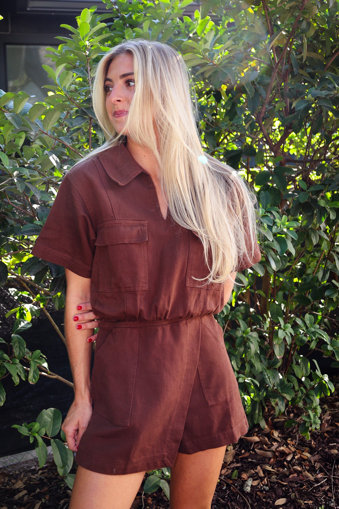 Woman wearing a brown jumpsuit standing in front of green foliage