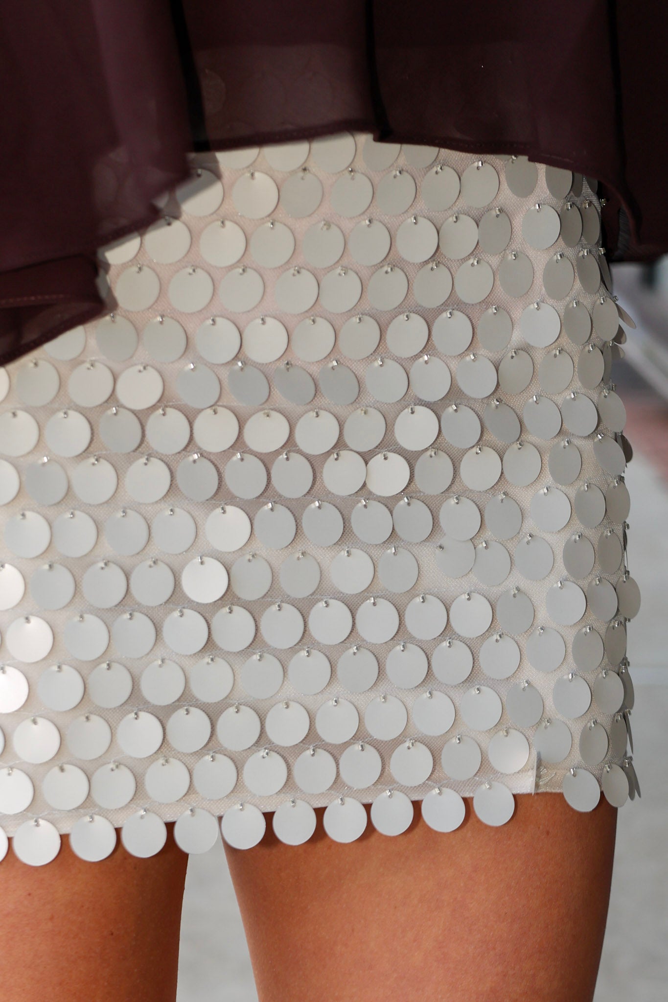A close-up of a white beige sequined mini skirt covered in overlapping round silver discs, worn with a dark sheer top.