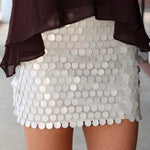A close-up of a person wearing a short white beige sequin mini skirt with a scalloped edge and a dark brown sheer draped top.