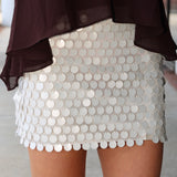 A close-up of a person wearing a short white beige sequin mini skirt with a scalloped edge and a dark brown sheer draped top.