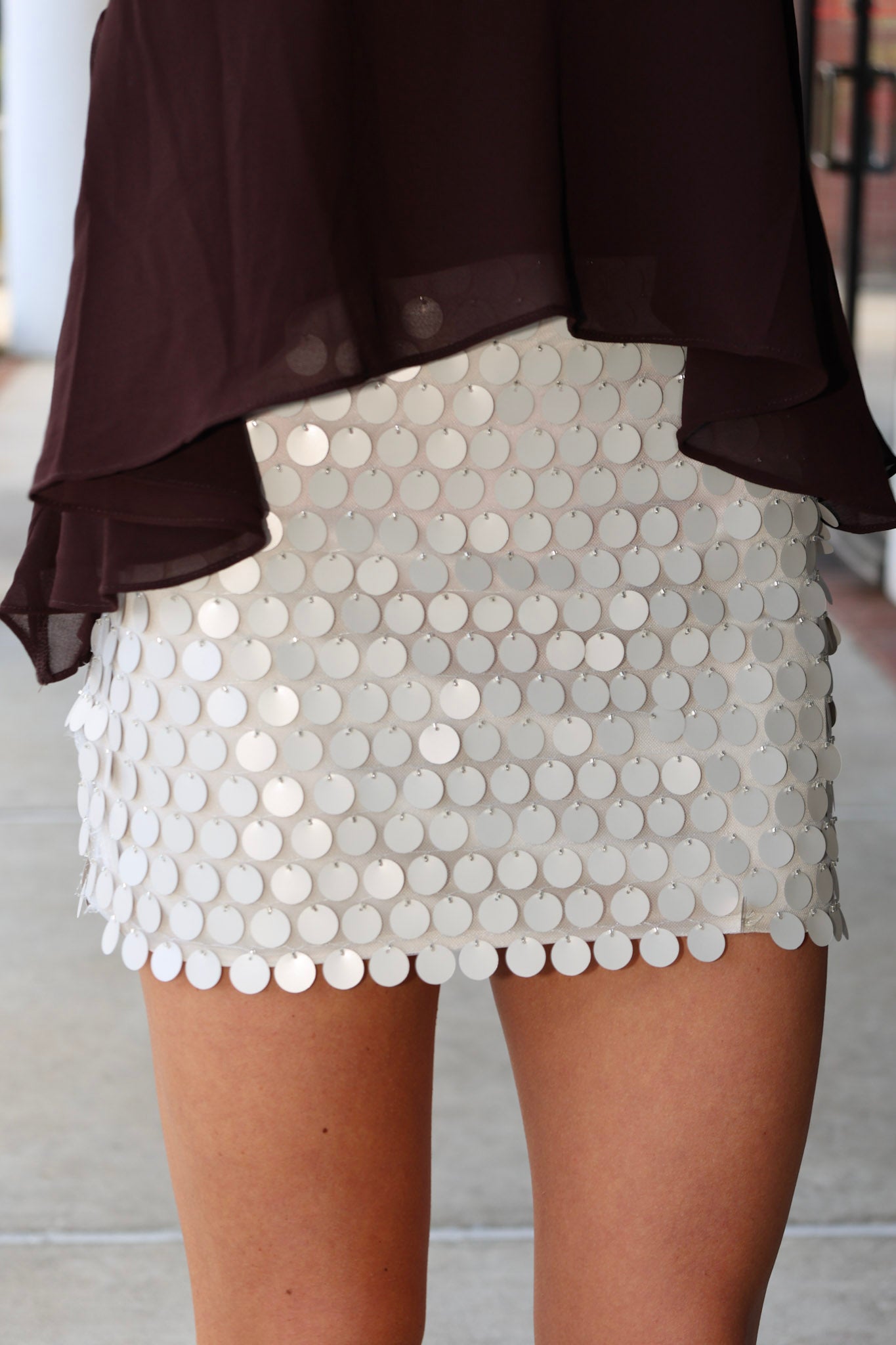 A close-up of a person wearing a short white beige sequin mini skirt with a scalloped edge and a dark brown sheer draped top.