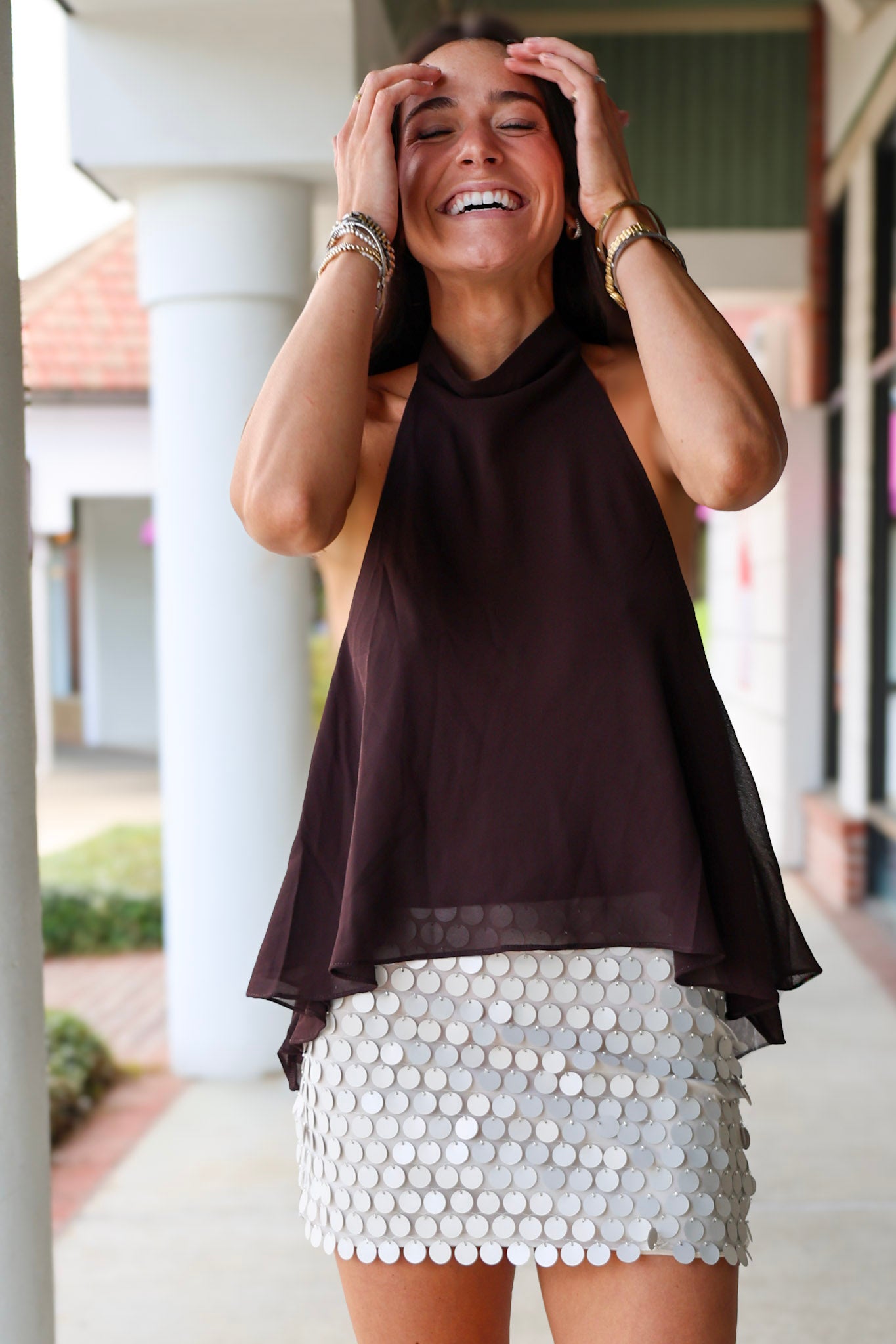 A woman laughing at Rodéo Boutique, wearing a dark brown sheer halter top and a white sequined mini skirt.