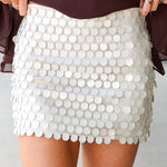A close-up of a person wearing a short white beige mini skirt covered in large round sequins, paired with a dark brown top. The person's hands are holding the sides of the skirt to show its shape.