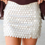 A close-up of a person wearing a short white beige mini skirt covered in large round sequins, paired with a dark brown top. The person's hands are holding the sides of the skirt to show its shape.