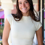 A smiling woman with long dark brown hair wearing a cream ribbed short-sleeve mock neck top and blue floral print jeans, standing in front of a softly blurred storefront background.