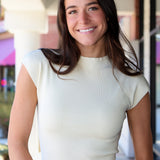 A smiling woman with long dark brown hair wearing a cream ribbed short-sleeve mock neck top and blue floral print jeans, standing in front of a softly blurred storefront background.