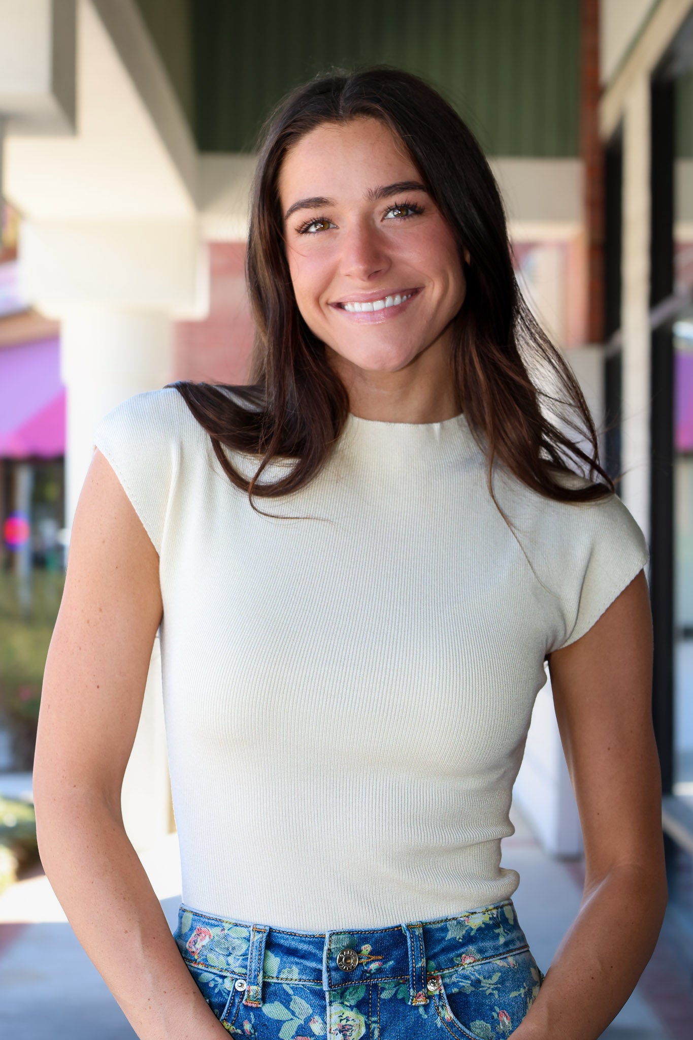 A smiling woman with long dark brown hair wearing a cream ribbed short-sleeve mock neck top and blue floral print jeans, standing in front of a softly blurred storefront background.