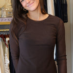 A woman wearing a dark brown long-sleeve crewneck top and blue jeans, standing in a clothing store with racks of clothes in the background.