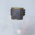 A square face ring featuring a gold-toned bezel-set pavé band with clear stones, flanked by twisted silver-toned side bands, displayed on a reflective white surface.